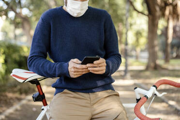 Mann mit Gesichtsschutz, der sich auf ein Fahrrad stützt, während er auf der Straße ein Mobiltelefon benutzt - AMPF00083