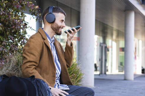 Geschäftsmann mit Kopfhörern, der mit seinem Mobiltelefon spricht, während er mit einer Tasche im Freien sitzt - PNAF00783