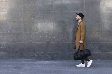 Fashionable businessman holding bag while walking against wall - PNAF00787