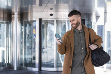 Lächelnder Geschäftsmann mit Tasche, der ein Mobiltelefon benutzt, während er vor einem Gebäude steht - PNAF00793