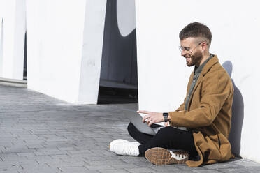 Lächelnder Geschäftsmann mit Jacke, der auf einem Gehweg sitzend an einem Laptop arbeitet - PNAF00800