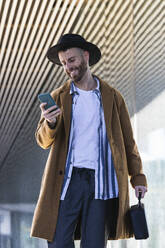 Mittlerer erwachsener Geschäftsmann mit Tasche, der im Freien stehend ein Mobiltelefon benutzt - PNAF00802
