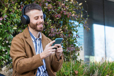 Smiling businessman wearing headphones using smart phone while sitting outdoors - PNAF00803