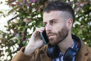 Mittlerer erwachsener Geschäftsmann mit Kopfhörern, der im Freien stehend mit seinem Mobiltelefon spricht - PNAF00804