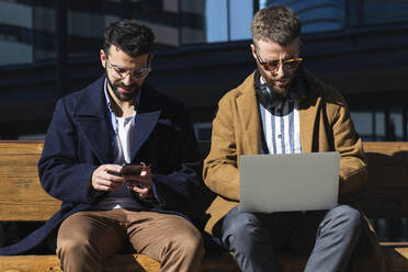 Young businessman using mobile phone while sitting by coworker working on laptop outdoors - PNAF00807