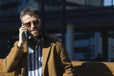 Geschäftsmann mit Sonnenbrille, der ein Mobiltelefon benutzt, während er auf einer Bank sitzt - PNAF00808