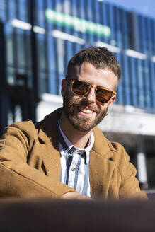 Mid adult businessman smiling while sitting outdoors during sunny day - PNAF00811