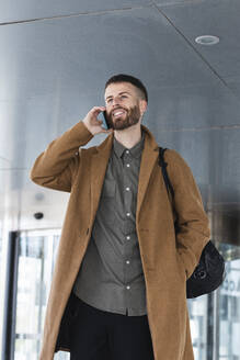 Männlicher Unternehmer, der im Stehen mit den Händen in den Taschen im Freien mit einem Mobiltelefon spricht - PNAF00817