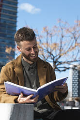 Lächelnder Geschäftsmann, der ein Dokument liest, während er im Freien sitzt - PNAF00825