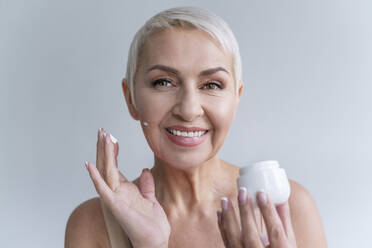 Lächelnde Frau trägt Gesichtscreme auf das Gesicht auf, während sie vor einem grauen Hintergrund steht - OIPF00384