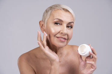 Ältere Frau trägt Gesichtscreme auf, während sie vor einem grauen Hintergrund steht - OIPF00386