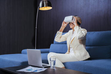 Lächelnde Geschäftsfrau, die ein Virtual-Reality-Headset benutzt, während sie mit einem Laptop auf dem Sofa sitzt - OIPF00412