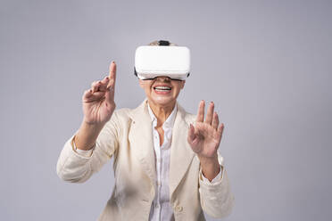 Senior businesswoman gesturing while using virtual reality headset while standing against gray background - OIPF00417