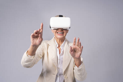 Ältere Geschäftsfrau gestikuliert, während sie ein Virtual-Reality-Headset benutzt und vor einem grauen Hintergrund steht - OIPF00417