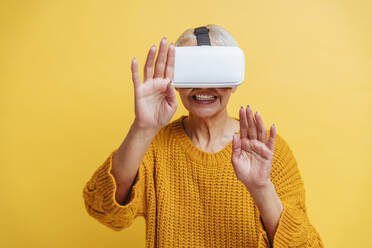 Smiling woman wearing virtual reality headset stretching hand while standing against yellow background - OIPF00422