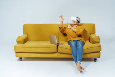 Senior woman with laptop using virtual reality headset while sitting on sofa - OIPF00429