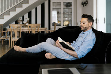 Mittlerer erwachsener Mann liest ein Buch, während er zu Hause auf dem Sofa sitzt - EGAF01930