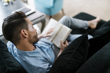 Mann liest ein Buch, während er zu Hause auf dem Sofa sitzt - EGAF01931
