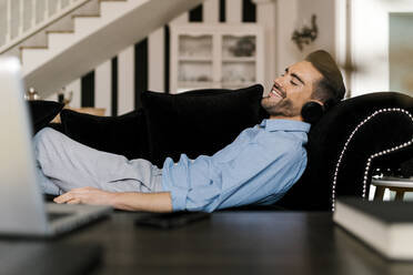 Smiling man wearing headphones relaxing on sofa at home - EGAF01937