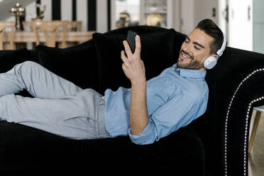 Smiling man with headphones using mobile phone while resting on sofa at home - EGAF01938