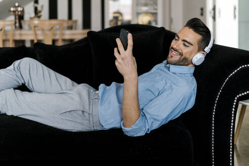 Lächelnder Mann mit Kopfhörern, der ein Mobiltelefon benutzt, während er sich zu Hause auf dem Sofa ausruht - EGAF01938