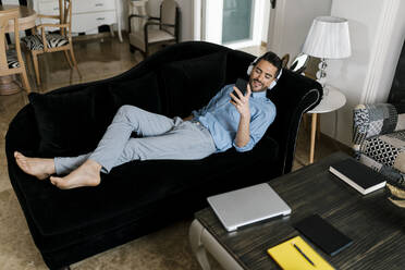 Smiling man with headphones using smart phone while relaxing on sofa at home - EGAF01939