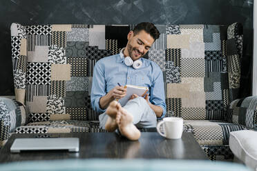 Mid adult businessman writing in book while sitting on sofa - EGAF01943