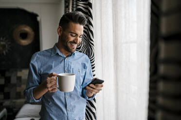 Lächelnder mittelgroßer erwachsener Mann mit Kaffeetasse, der ein Mobiltelefon benutzt, während er zu Hause steht - EGAF01949