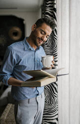 Mann mit Kaffeetasse beim Lesen eines Buches im Stehen zu Hause - EGAF01950