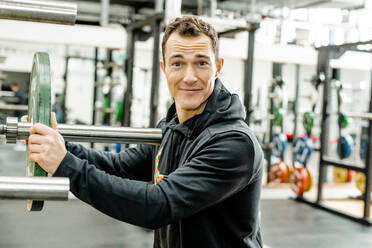Lächelnder männlicher Sportler in Sportkleidung, der in einem Fitnessstudio eine Gewichtsscheibe hält - KVF00179