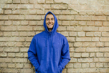 Smiling male athlete in blue jacket against brick wall - KVF00190