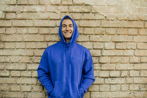Lächelnder männlicher Sportler in blauer Jacke vor einer Backsteinmauer - KVF00190