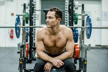 Shirtless male athlete looking away while sitting at bench press in gym - KVF00193