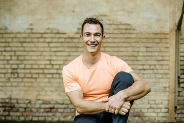 Lächelnder männlicher Sportler, der in einem Fitnessstudio an einer Mauer sitzt - KVF00195