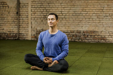 Male athlete with cross legged meditating against brick wall in health club - KVF00198