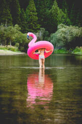 Girl wading through river and carrying flamingo floatie - CAVF93544