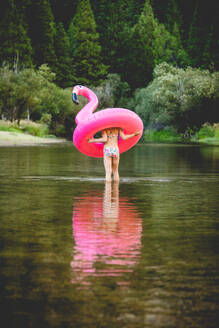 Mädchen watet durch einen Fluss und trägt ein Flamingo-Schwimmteil - CAVF93544