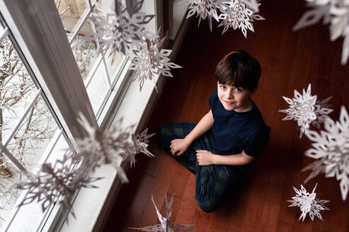 Junge sitzt auf dem Boden neben dem Fenster, umgeben von Papierschneeflocken. - CAVF93552