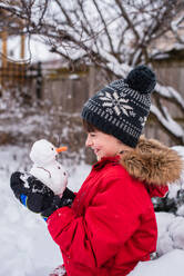 Junge hält an einem verschneiten Tag im Freien einen kleinen Schneemann in den Händen. - CAVF93555
