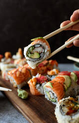 Nahaufnahme von Stäbchen, die eine Sushi-Rolle über einem Teller mit Lebensmitteln halten. - CAVF93561