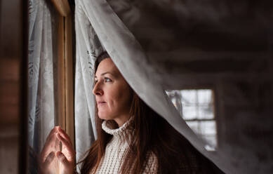 Attraktive Frau schaut durch ein Fenster hinter einem Spitzencuratin. - CAVF93564