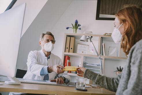 Male doctor wearing protective face mask giving vaccination certificate to patient while sitting at office - MFF07381