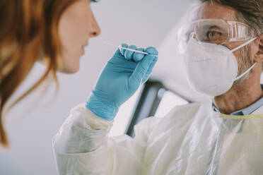Männlicher Arzt mit Schutzanzug und Gesichtsmaske, der einen Nasenabstrich bei einem Patienten im Untersuchungsraum vornimmt - MFF07391