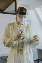 Ärztin in Arbeitsschutzkleidung, die im Untersuchungsraum stehend eine Probe in einen Plastikbeutel gibt - MFF07407