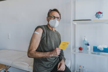 Male patient wearing protective face mask with bandage on arm holding vaccination certificate while sitting at examination room - MFF07439