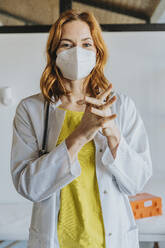 Weiblicher Arzt im mittleren Erwachsenenalter mit Gesichtsmaske desinfiziert seine Hände mit Desinfektionsmittel, während er in einer Klinik steht - MFF07463