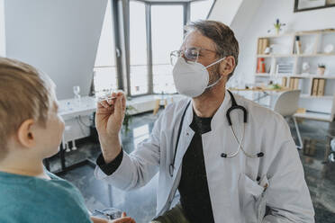 Männlich, mit Gesichtsschutzmaske, Kinderarzt, der einen Nasenabstrich bei einem Jungen macht, während er in der Klinik steht - MFF07471