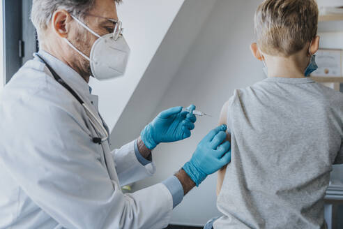 Arzt mit Gesichtsschutz, der einem Jungen den Impfstoff COVID-19 verabreicht, während er in einer Klinik steht - MFF07479
