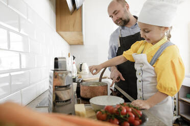 Smiling daughter wearing apron and chef's hat cooking food while standing by father in kitchen - KMKF01533