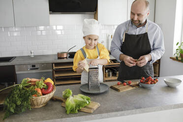 Mädchen mit Reibe bei der Zubereitung von Essen mit Vater in der Küche zu Hause stehend - KMKF01535